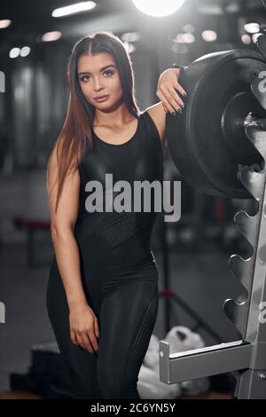Bella affascinante modello femminile che si posa in un moderno studio fitness vicino alla fila di barbell, guardando dritto con piacevole espressione, ritratto, vista ravvicinata Foto Stock