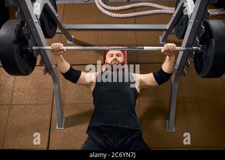 Ritratto di brutale uomo maleducato con barba spessa e testa calva, sdraiato su panchina con mani sollevate, reggendo la barra di ferro con passione, respiro, ottenere st Foto Stock