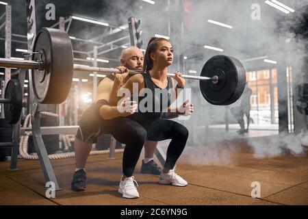 Donna sportiva cool attraente vestita di sportswear nero, squatting con il barbell pesante, prendendo l'aiuto di allenatore professionale di fitness, l'addestramento Foto Stock