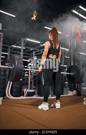 Forte alto bodybuilder femminile che si prepara a sollevare pesante barbell, in piedi con la schiena alla macchina fotografica, praticando con barbell in palestra sala, shot dal basso, Foto Stock