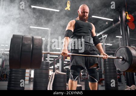 Uomo sportivo attivo con barba spessa, vestito di abbigliamento sportivo nero, pratica in garage sotterraneo, guardando giù esprimendo potere, allenandosi con barbel Foto Stock