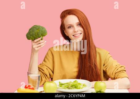 giovane donna allegra con lunghi capelli di zenzero vestito in felpa gialla con cibo vegetariano con broccoli, su sfondo rosa. Salute Foto Stock