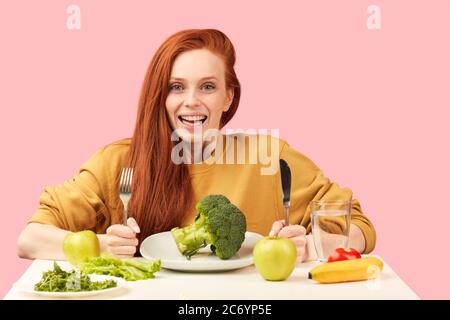 Studio ritratto di giovane allegra bella donna con lunghi capelli di zenzero vestito in felpa gialla con cibo vegetariano insieme con broccoli, contro rosa Foto Stock