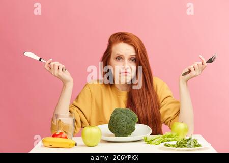 Una giovane donna dai capelli rossi, incerta e perplessa, seduta a tavola, è servita con broccoli che si infrangono con spalle e guarda la telecamera con uno sguardo interrogatorio. Foto Stock