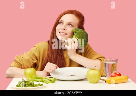 Donna snella e felice seduta a tavola con cibo vegano sano, tenendo in mano i broccoli freschi, guardando verso l'alto, sognando di torta gustosa, tentazione Foto Stock