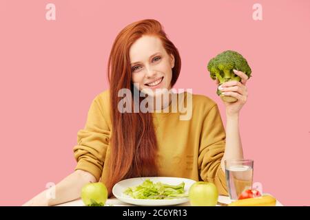 Giovane soddisfatto soddisfare donna sulla dieta mangiare piatto di verdure fresche avendo buon appetito e umore allegro, seduta isolata al tavolo sopra backg rosa Foto Stock