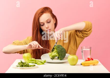 Studio ritratto di giovane allegra bella donna con lunghi capelli di zenzero vestito in felpa gialla con cibo vegetariano insieme con broccoli, contro rosa Foto Stock