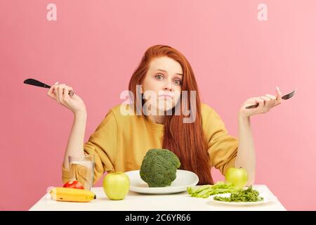 Una giovane donna dai capelli rossi, incerta e perplessa, seduta a tavola, è servita con broccoli che si infrangono con spalle e guarda la telecamera con uno sguardo interrogatorio Foto Stock