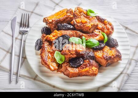 Ricetta di caduta di costolette di maiale con osso di bambino corto brasate con prugne, brodo, cipolla e aglio, decorate con basilico fresco, servite su un piatto bianco Foto Stock