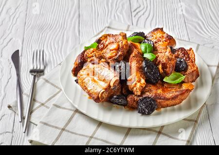 Stufato con prugne secche, cipolla e aglio di maiale, decorato con basilico fresco, servito su un piatto bianco su fondo di legno bianco con posate, Foto Stock