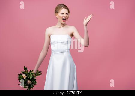 sposa arrabbiata in bianco abito da sposa con fiori e gesturing su rosa Foto Stock