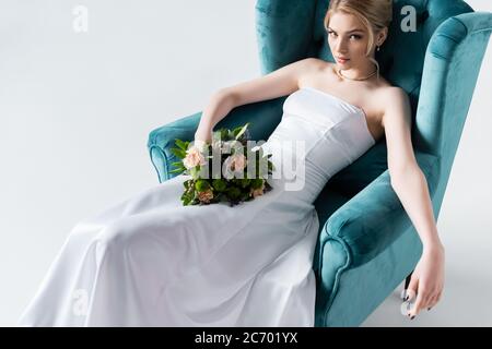 vista ad alto angolo della sposa in elegante abito da sposa con fiori mentre si siede in poltrona su bianco Foto Stock