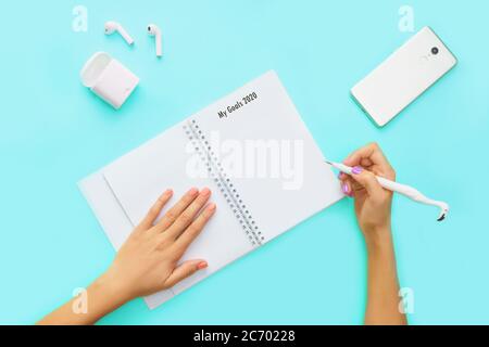 Concetto di spazio di lavoro minimo. Obiettivi 2020, elenco chek. Vista dall'alto di cuffie, telefono e notebook con mani su sfondo blu. Disposizione piatta. Foto Stock