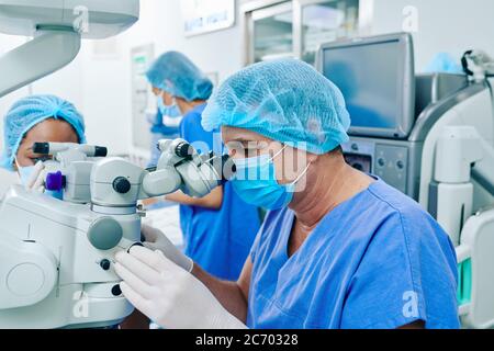Chirurgo maturo in guanti e maschera medica utilizzando apparecchiature moderne quando si esegue l'operazione di correzione della visione laser Foto Stock