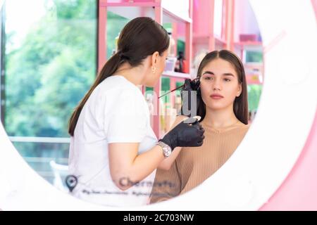 Il pittore di sopracciglia del maestro di bellezza dipinge le sopracciglia colorandole con hennè. Donna fa un make-up lavorando in guanti neri in un salone di bellezza. Concetto di fronte Foto Stock