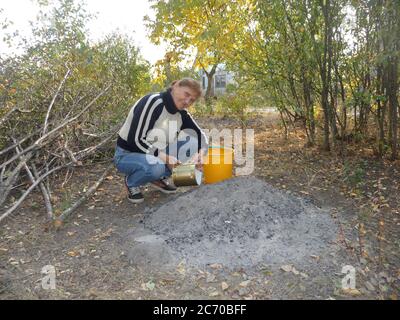 Primo piano ritratto di una donna anziana sorridente che sta sbattendo in cortile vicino a un mucchio di cenere di legno. Foto Stock