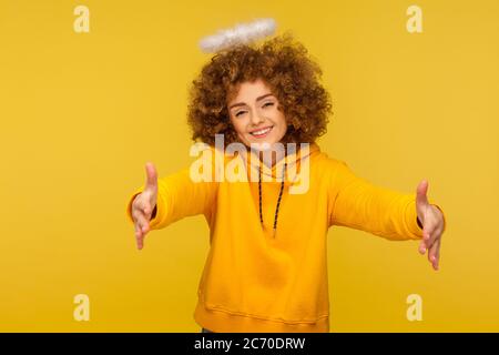 Ritratto di generosa donna hipster con i capelli ricci e il cuore gentile con san nimbus che alza le mani per abbracciare, dando libero abbracci e sorridendo con amore. Indoo Foto Stock