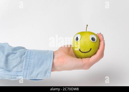 La mano tiene verde mela-smiley con gli occhi di Googly. Divertente Apple su sfondo bianco. Foto Stock