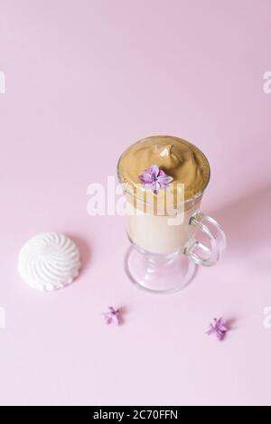 Una bella bevanda a base di caffè dalgona. Tazza di caffè in vetro trasparente con schiuma e fiore lilla su sfondo lilla. Dolcezza alla marshmallow-bevanda Foto Stock