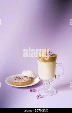 Bella dalgona bere un caffè di schiuma in una tazza trasparente e fiori di lilla su uno sfondo viola. Dolci su un piatto: Donat e tenero marshmal Foto Stock