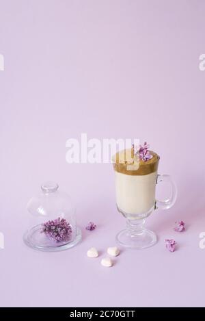 Caffè dalgona in tazza trasparente con tubo di carta, un piatto con fiori lilla e dolci su fondo lilla con uno spazio per la copia Foto Stock