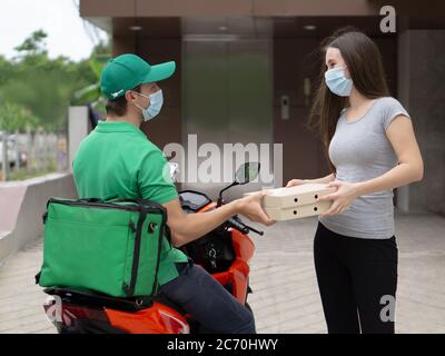 Concetto di consegna di cibo e pacchi. Il personale addetto alla consegna in uniforme verde su una motocicletta ha consegnato la scatola per la pizza al giovane cliente. Entrambi indossano un viso Foto Stock