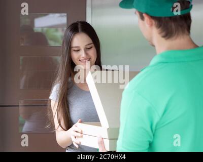 Concetto di consegna di cibo e pacchi. Il personale addetto alla consegna in uniforme verde ha consegnato la scatola per la pizza al giovane cliente. La donna sorrise felice Foto Stock