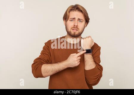 Guarda ora! Ritratto di un uomo impaziente preoccupato con barba in felpa che punta orologio da polso e che guarda ansioso infelice, mostrando orologio per affrettarsi. Foto Stock