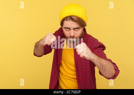 Aggressivo hipster con il cappello di berretto, camicia a scacchi che tiene pugni aggrappati pronti a pugilato, pugilato con espressione sicura, combattimento Foto Stock