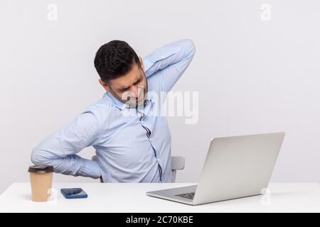 Uomo d'affari stanco seduto in ufficio con un computer portatile, si sente sfinito di mal di schiena, massaggiare i muscoli rigidi per alleviare gli spasmi, sedentario vita Foto Stock