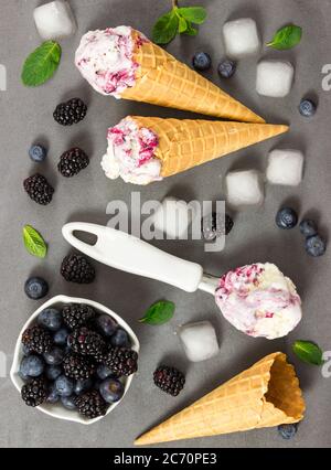 Gelato alla frutta con mora fresca e mirtillo, cubetti di ghiaccio e cono gelato vuoto su fondo di pietra grigia. Vista dall'alto. Foto Stock