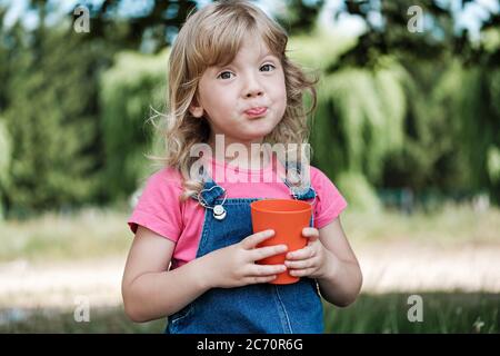 Carina bambina bionda pursing le labbra come lei beve succo da una tazza rossa colorata all'aperto in giardino in estate in primo piano Foto Stock