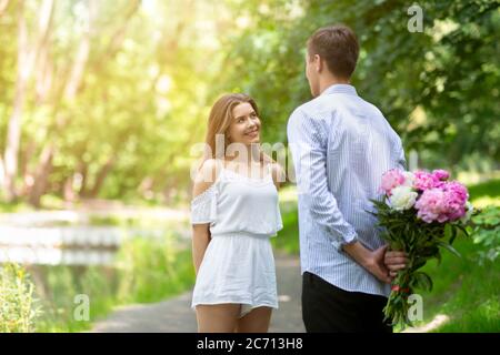 Giovane uomo che si congratula con la ragazza per il compleanno, nascondendo bouquet di fiori dietro la schiena al parco. Spazio vuoto Foto Stock