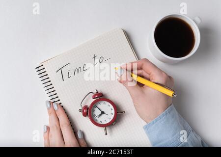 La mano della donna scrive nel taccuino il tempo di iscrizione a. Blocco note e sveglia vintage. Pianificazione e caffè. Foto Stock