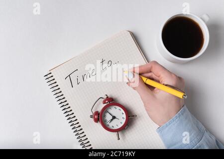 La mano della donna scrive nel taccuino il tempo di iscrizione a. Blocco note e sveglia vintage. Pianificazione e caffè. Foto Stock