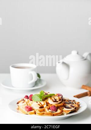Cialde belghe con lamponi, banana, sciroppo di cioccolato. Colazione con tè su sfondo bianco, vista laterale, verticale Foto Stock