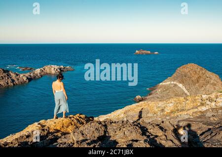 giovane donna determinata in piedi sulla cima di scogliera rocciosa sul mare al tramonto, riflettendo e meditando nel vento soffiato Foto Stock