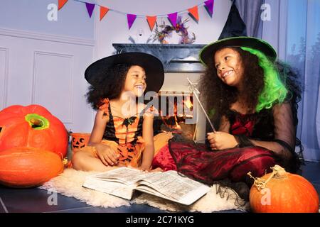 Due ragazze sorridenti felici leggono il libro di mago in costumi di Halloween strega gettano un incantesimo con wand magico che tiene seduto vicino al camino Foto Stock