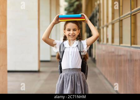Ritorno a scuola. Scolaretta in uniforme che tiene un mucchio di libri sulla sua testa vicino alla costruzione della scuola. Foto Stock