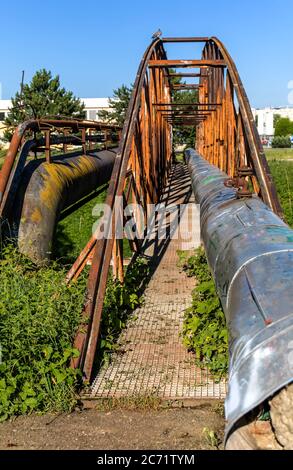 Vecchio ponte di ferro arrugginito sul fiume con un tubo di vapore. Alimentazione termica. Apparecchiature industriali. Foto Stock