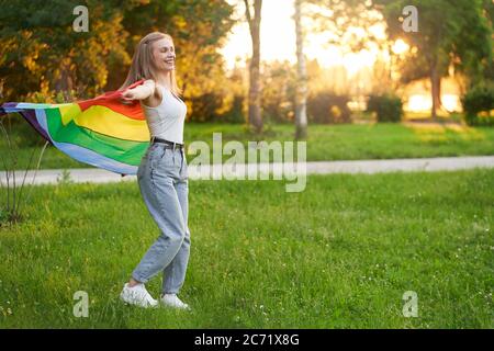 Vista laterale di felice ridendo ragazza dacing, arcobaleno oscillante bandiera lgbt sul vento dietro la schiena, tramonto estivo sullo sfondo. Bella giovane donna che si diverte nel parco. Concetto di tolleranza delle minoranze lgbt. Foto Stock