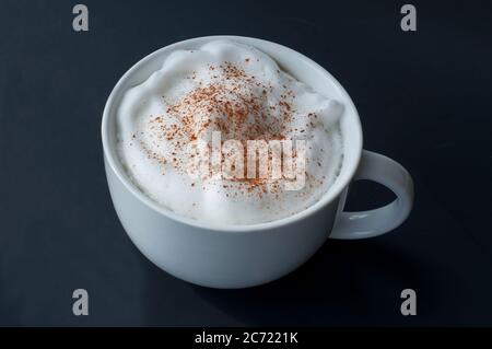 Il latte di caffè con la schiuma di latte e la cannella si cospargano in una grande tazza bianca su uno sfondo scuro. Questa bevanda calda è perfetta per la mattina e quando serve Foto Stock