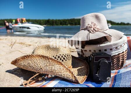 Cesto in vimini, cappelli estivi. Una macchina fotografica sulla sabbia. Su una barca di sfondo sfocato. Alla spiaggia della Finlandia. Foto di alta qualità Foto Stock