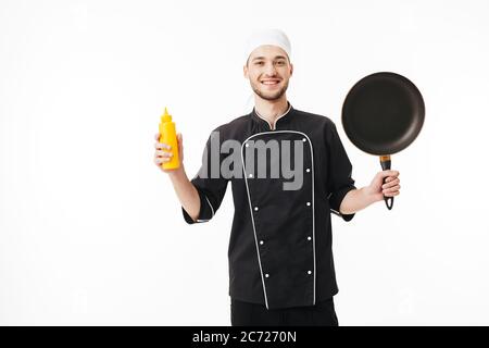 Giovane chef sorridente in uniforme nera che tiene in mano una padella e una bottiglia di senape che guarda felicemente in macchina fotografica su sfondo bianco Foto Stock