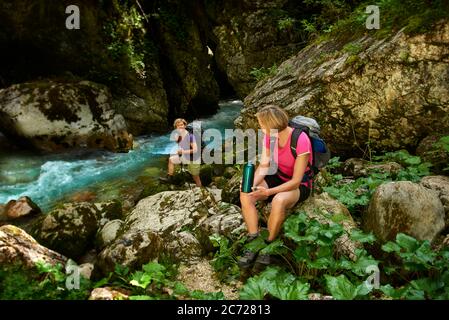 Due donne riposano, bevono acqua, godono la vista e parlano in un canyon vicino al fiume di montagna. Distanza di sicurezza, zaino, natura. Foto Stock