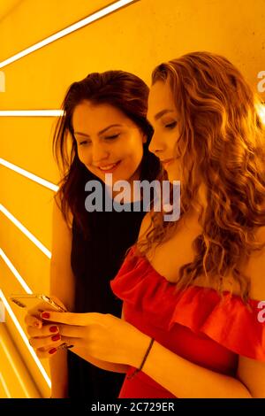 Due amiche che guardano lo schermo di uno smartphone sorridente. Carina ragazza bionda dai capelli lunghi con un vestito rosso festivo. Donna brunetta con abito nero. Foto Stock