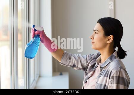 Giovane donna in guanti per la pulizia della finestra con detergente spray a casa Foto Stock