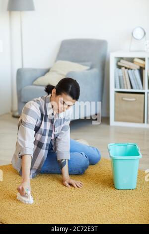 Giovane donna seduta sul pavimento e lavando il tappeto con detergente in soggiorno Foto Stock
