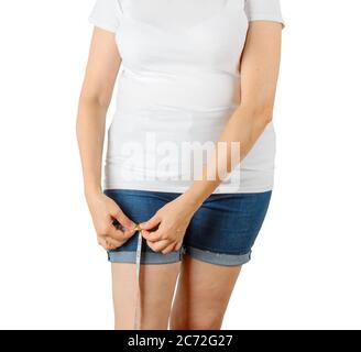 La donna adulta caucasica matura inizia ad aumentare il peso e misura la coscia con un nastro di misurazione. Donna vestita con una T-shirt bianca e un corto in denim blu Foto Stock