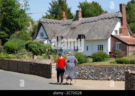 Scena di strada del villaggio inglese in estate; due donne che camminano su una strada di fronte ad un cottage tradizionale con tetto di paglia, Dalham Village, Suffolk Inghilterra UK Foto Stock